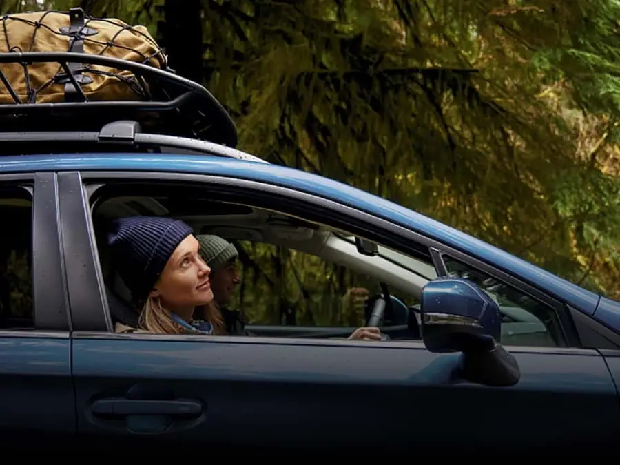 Couple driving blue Subaru through a park with the windows down. 