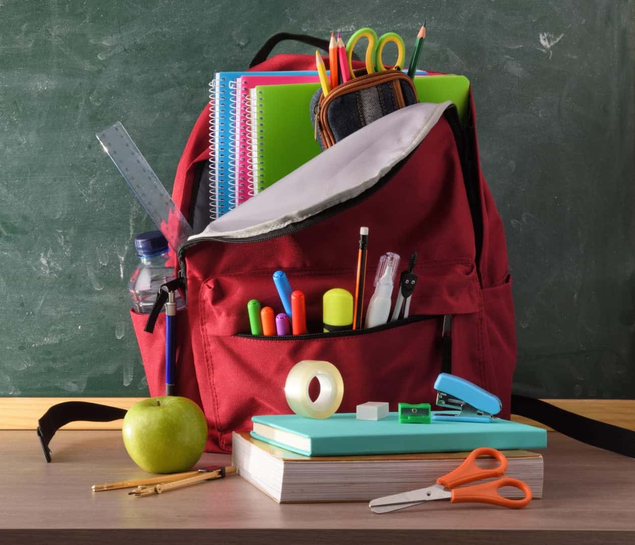 Red backpack filled with notebooks, pencils, scissors, markers, and other school supplies in front of a classroom chalkboard.