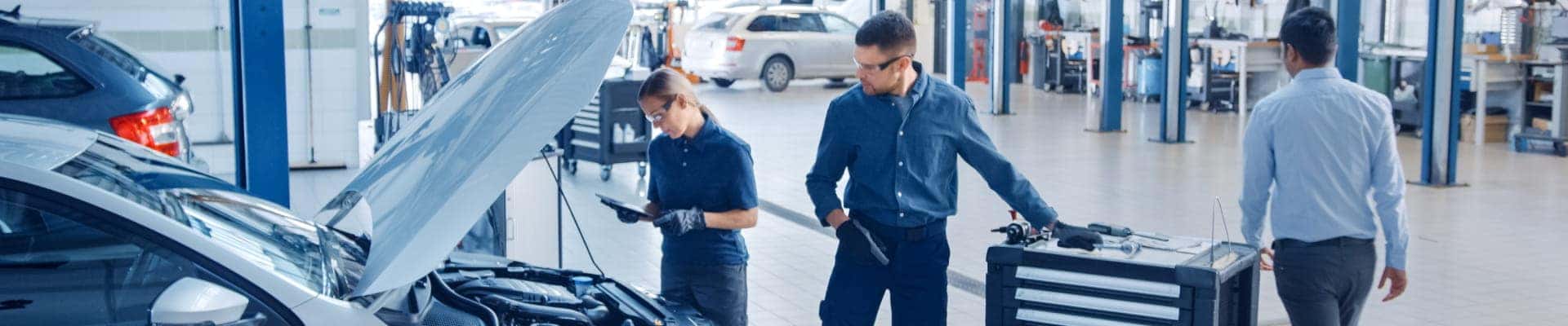 Automotive service technicians inspecting a vehicle with its hood open inside a service center.