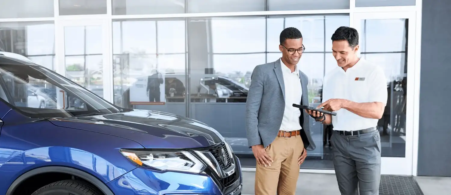 A Nissan finance consultant going through figures on a tablet with a customer beside a blue Nissan SUV in the showroom