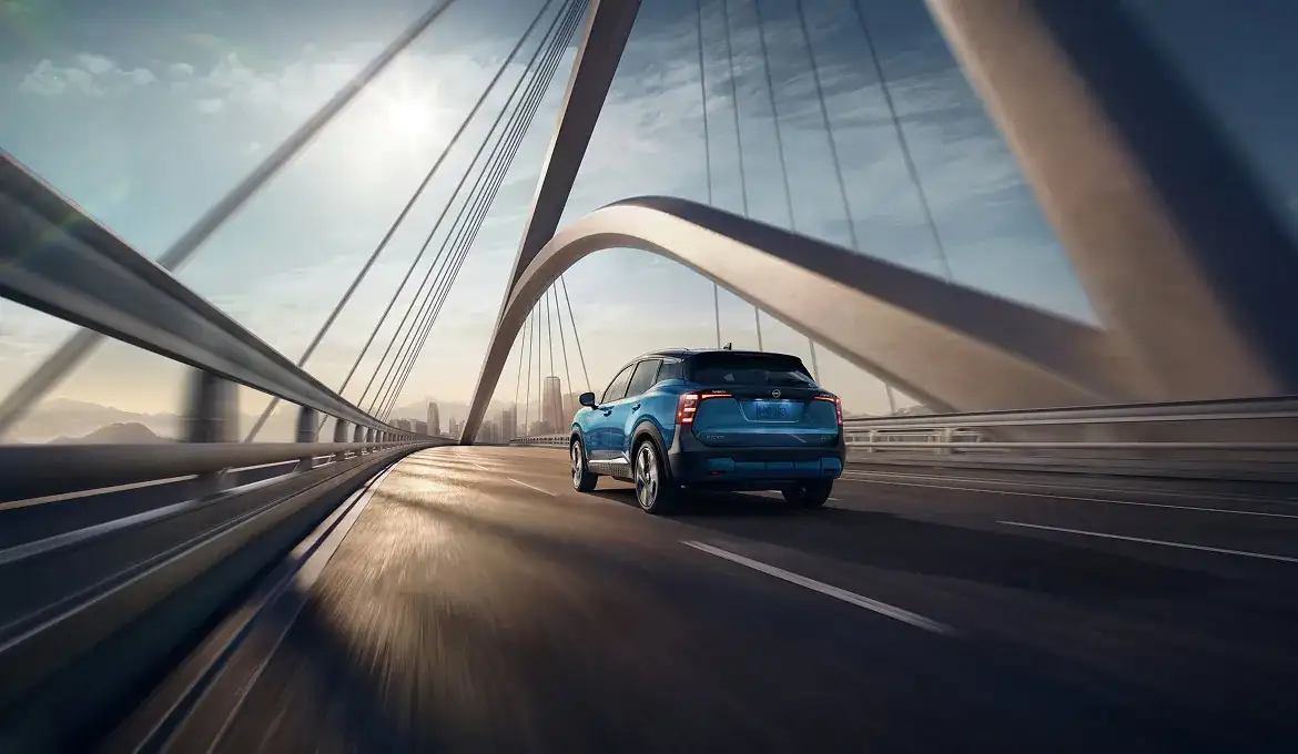A blue Nissan Kicks seen from behind, crossing a cable-stayed bridge toward a city skyline at sunrise