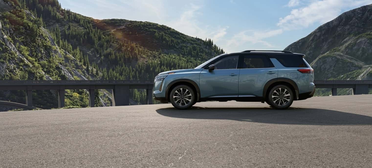 A 2026 Nissan Pathfinder in pale sage, side profile, on a mountain road with forested peaks behind