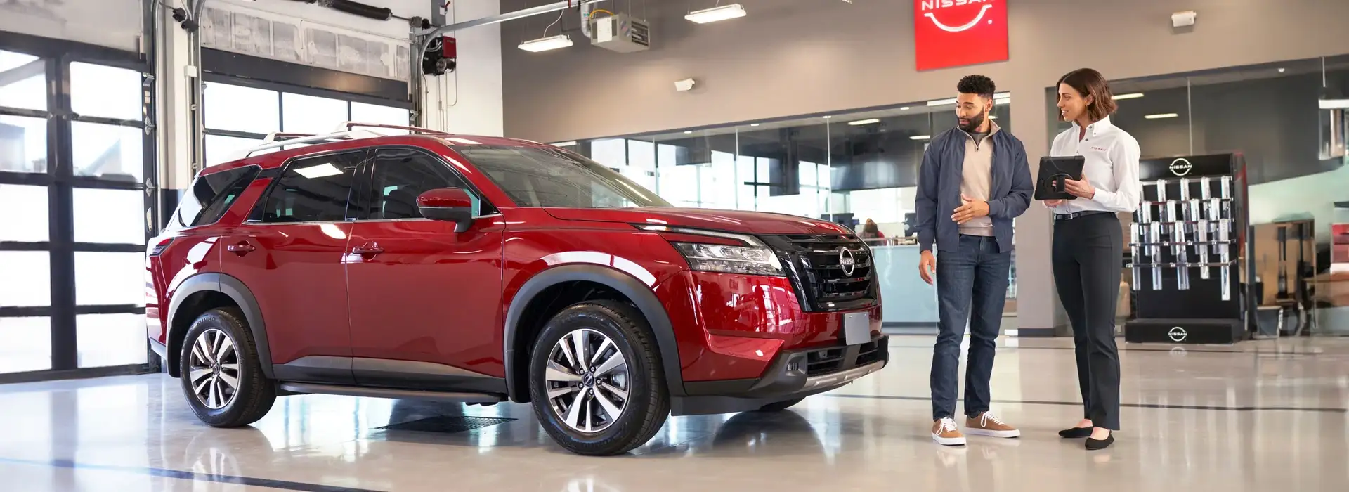 A Nissan service advisor with a tablet talking to a customer beside a red Nissan Pathfinder in the service reception bay