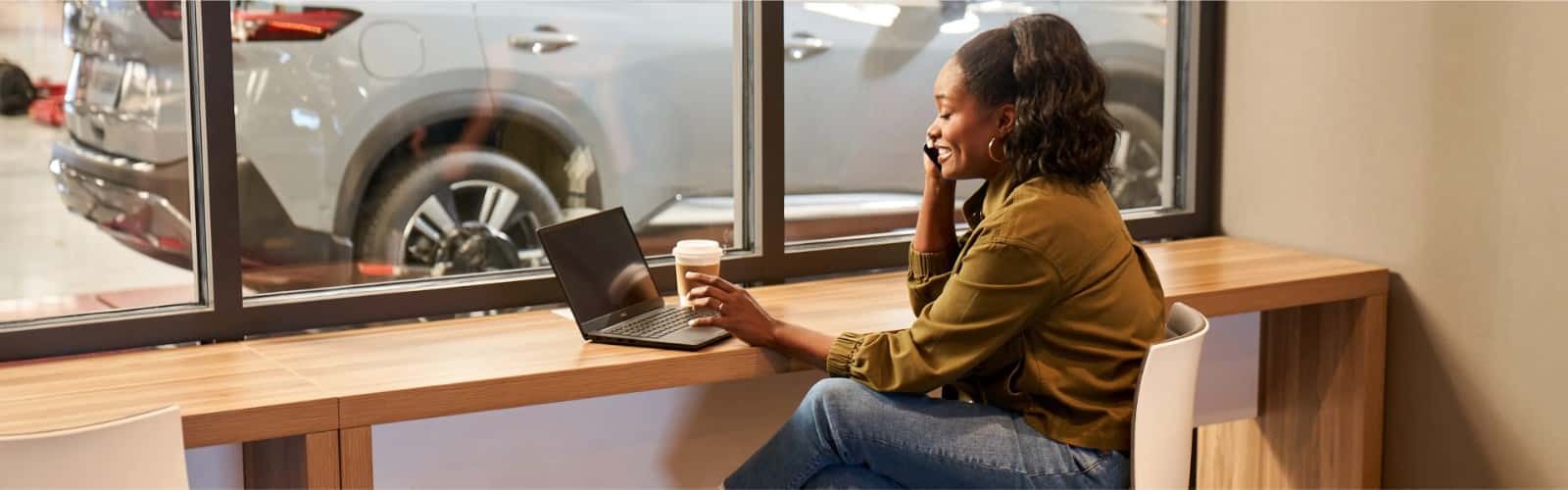 A customer working on a laptop at a counter in the service waiting lounge, a Nissan visible through the window