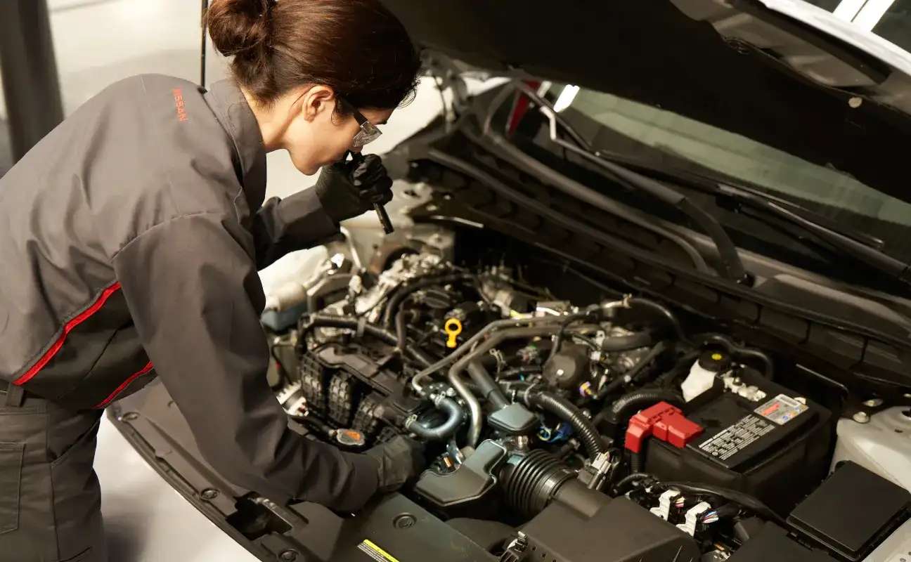A Nissan technician in safety glasses inspecting an engine bay with a flashlight