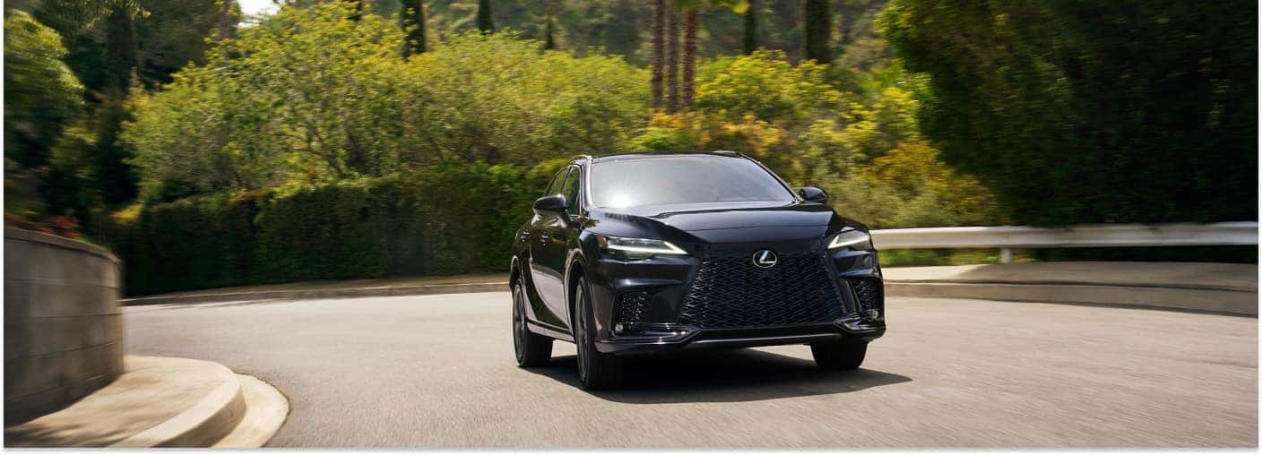 Front view of a black 2026 Lexus RX driving on a winding paved road lined with trees.