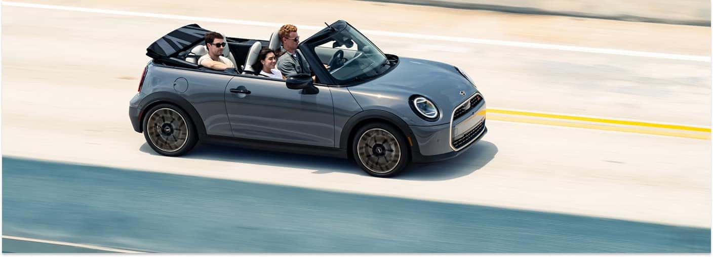 Side view of a grey 2026 MINI Convertible driving on a highway with the top down.