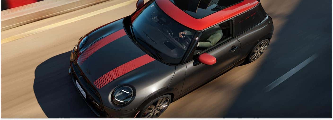 Dark grey 2026 MINI John Cooper Works Hardtop 2-Door with red roof and red hood stripes driving on a road.