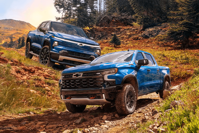  Two Chevrolet pickup trucks, a blue Silverado EV Trail Boss and a bright blue Silverado ZR2, are driving on a rugged, dirt trail in a mountainous, forested landscape.
