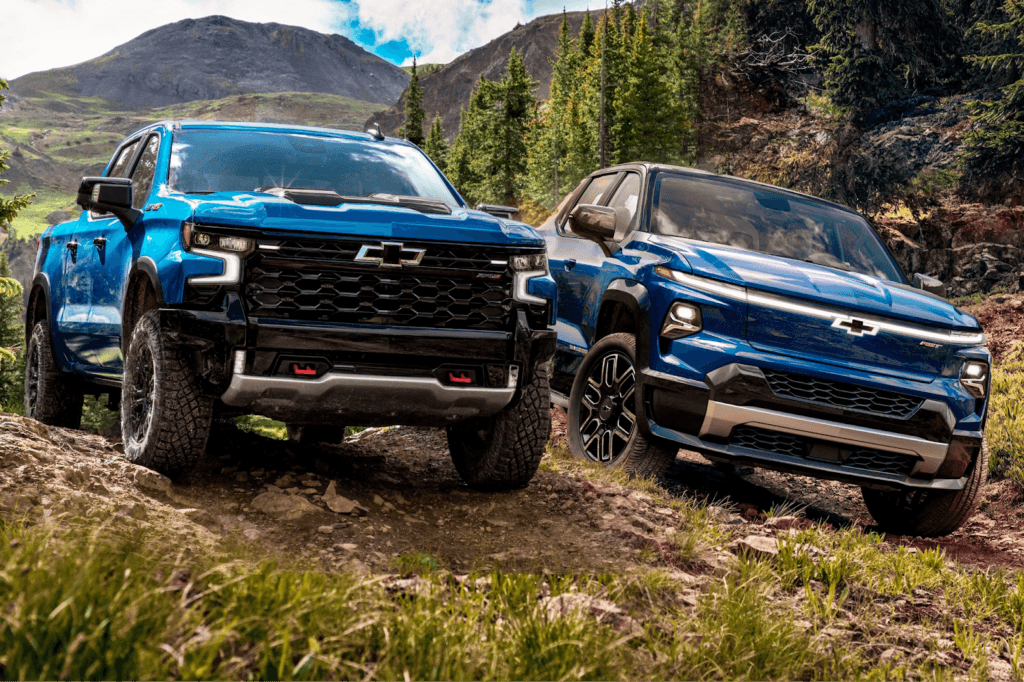  Two Chevrolet Silverado pickup trucks are shown driving off-road in a rocky, mountainous landscape with evergreen trees. The blue Silverado ZR2 is in the foreground, and a dark blue electric Silverado EV is slightly behind and to the right, highlighting both trucks' off-road capabilities and modern designs. 