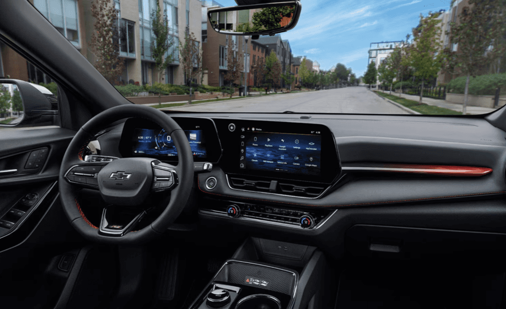 The dashboard and cockpit view of a vehicle, showing the digital instrument cluster and the large infotainment screen. The interior features a black color scheme with red stitching and accents, and the windshield view shows a residential street with row houses.