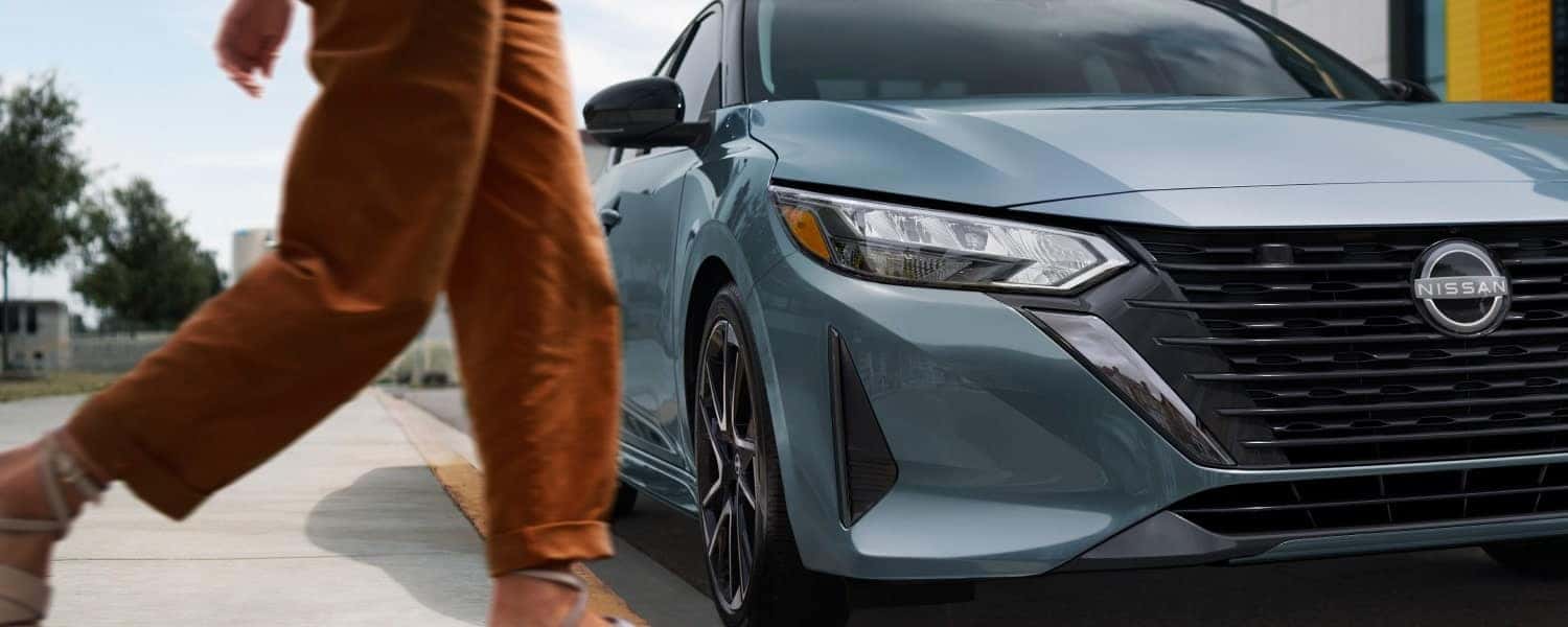 A blue Nissan Sentra parked by a curb with a person walking by