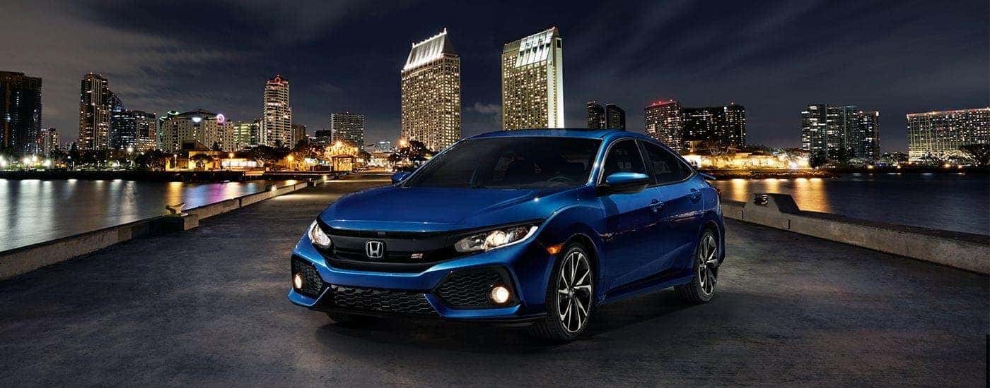 A blue 2017 Honda Civic Si parked in front of a cityscape at night.