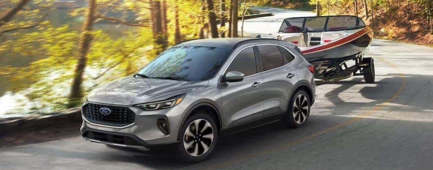A silver 2024 Ford Escape is shown towing a boat on a windy road.