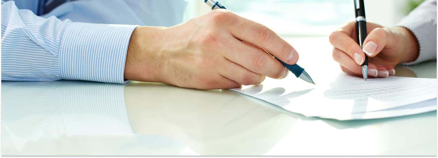 Close-up of two people filling out and signing auto financing paperwork at a desk with pens.