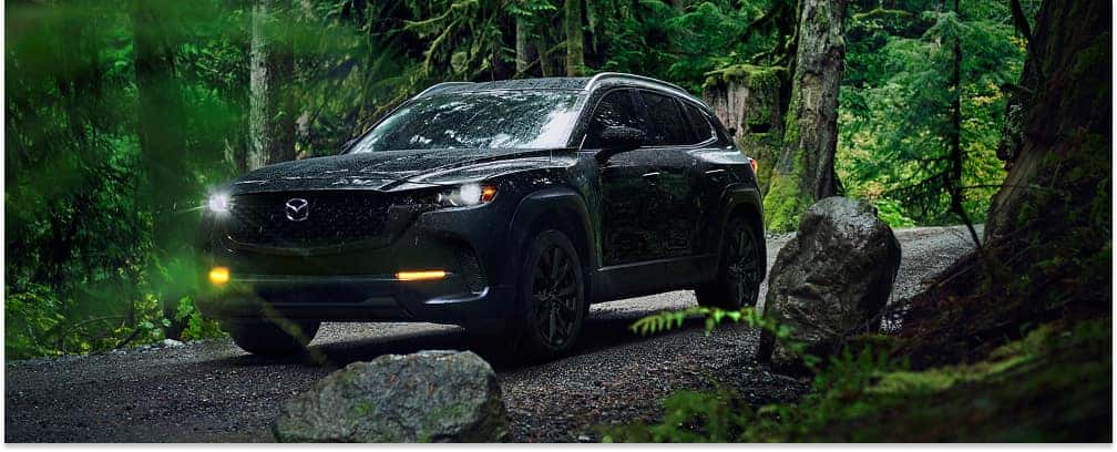 Front view of a black 2026 Mazda CX-50 driving on a wet dirt road through a forest with headlights illuminated.