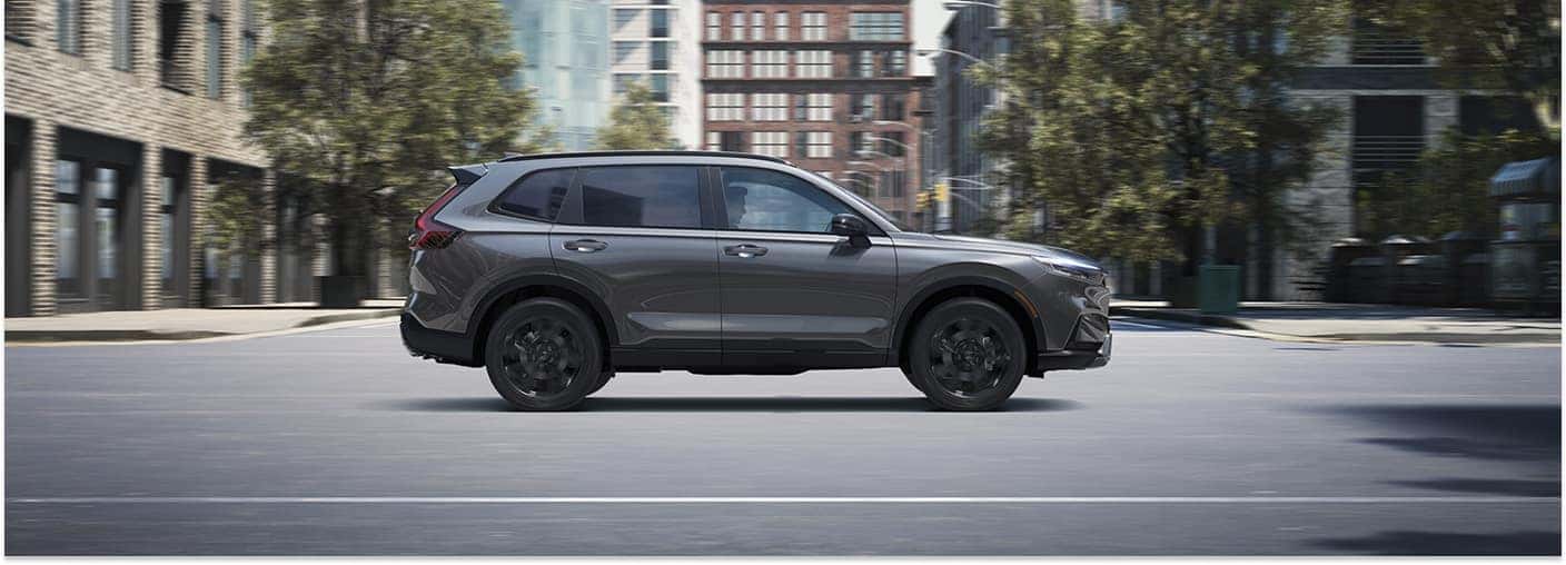 Side profile of a dark gray 2026 Honda CR-V SUV driving along an urban city street.