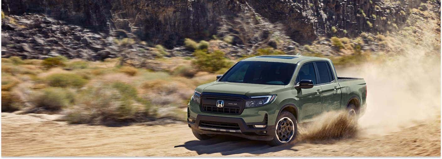 Front three-quarter view of an olive green 2026 Honda Ridgeline driving on a dusty dirt trail in a desert landscape.