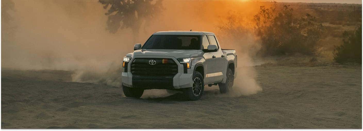 Front three-quarter view of a white 2026 Toyota Tundra driving off-road in a dusty desert setting at sunset.