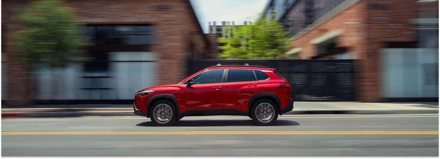 Red 2026 Toyota Corolla Cross driving past brick buildings in a side profile view.