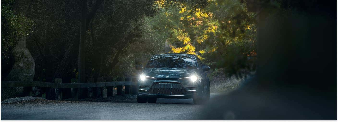 Dark gray 2026 Toyota Corolla driving on a shaded road with headlights illuminated, front view.