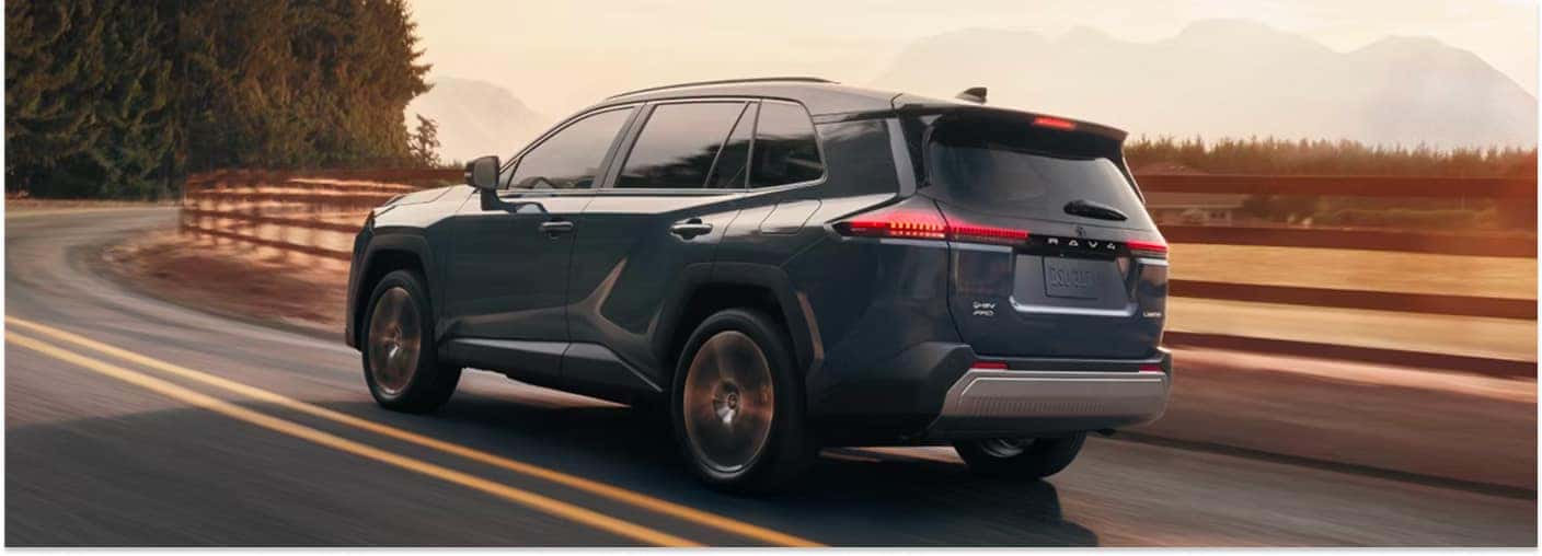 Dark gray 2026 Toyota RAV4 SUV driving on a paved road, rear three-quarter view.