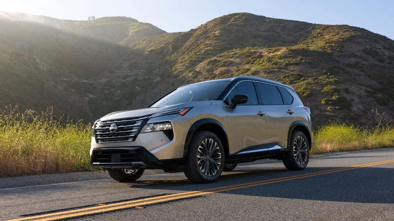 A silver Nissan Rogue parked on a two-lane road with sunlit hills and yellow wildflowers in the background.