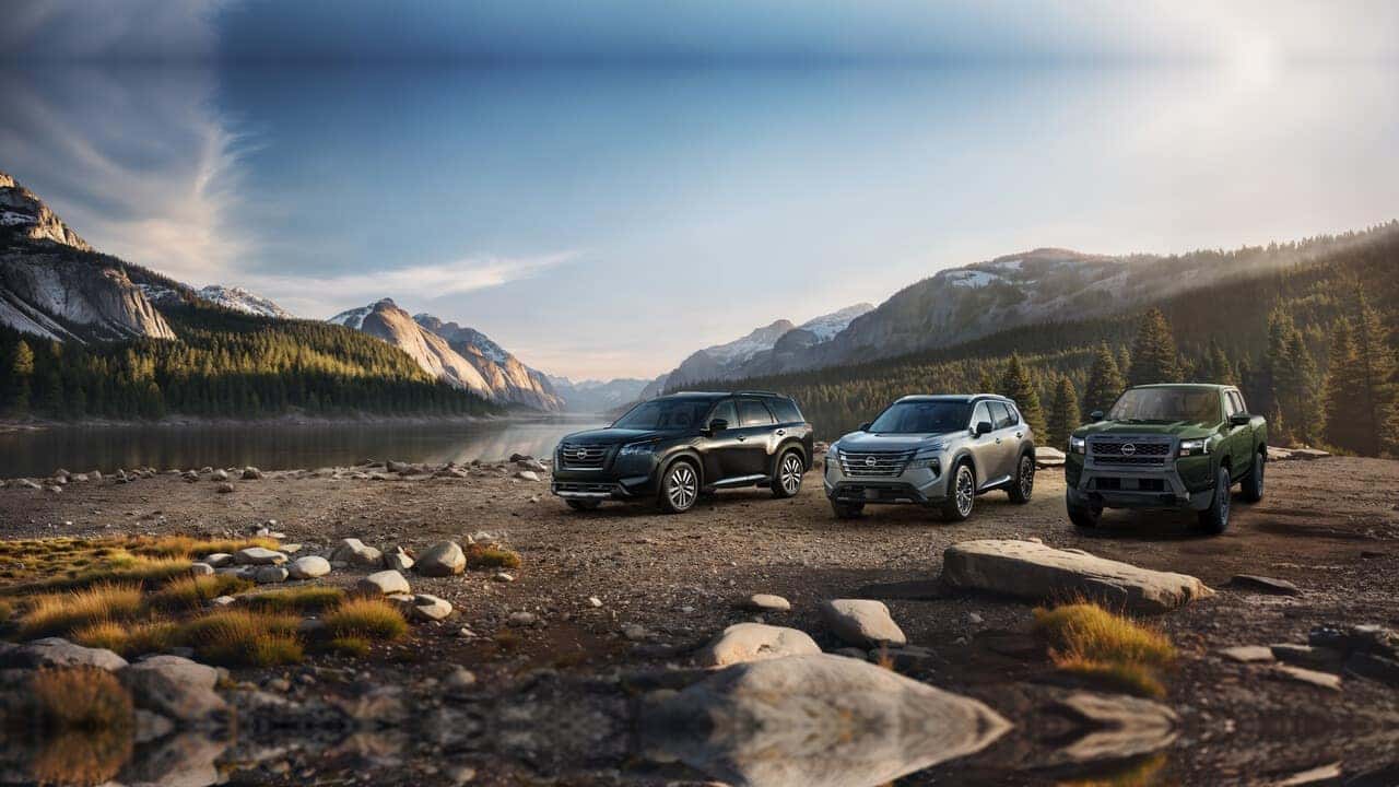 A Nissan Pathfinder, Rogue, and Frontier parked along a rocky lakeshore with forested mountains in the background.
