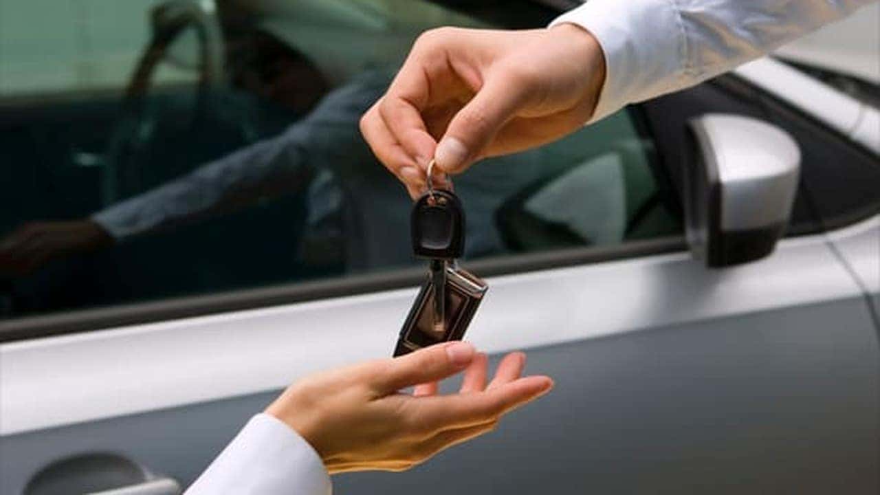 A person hands car keys to another person beside a silver car.
